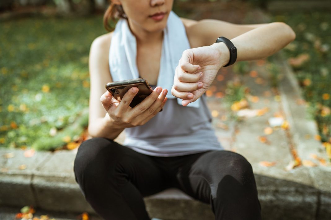 Anonymous female in sportswear using smartphone and smart watch after exercising on sports ground